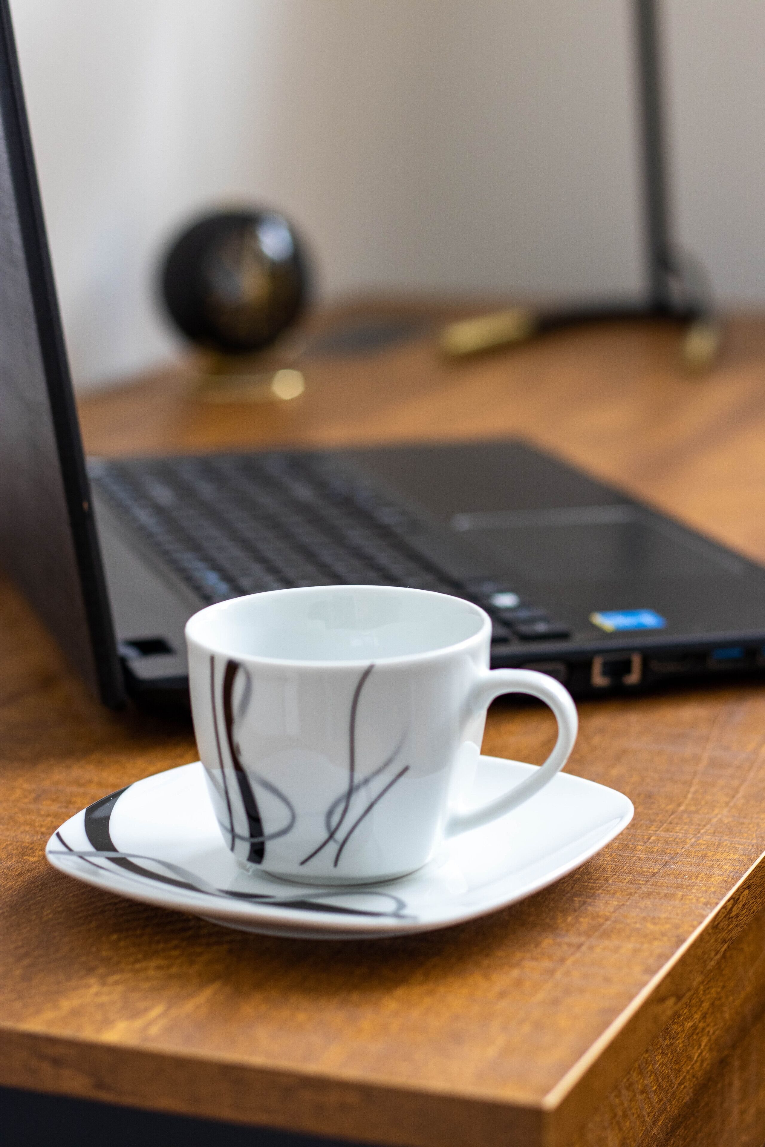 Een witte mok met zwarte designs op een schotel, geplaatst op een houten tafel, met een laptop en een zwart, bolvormig object op de achtergrond.