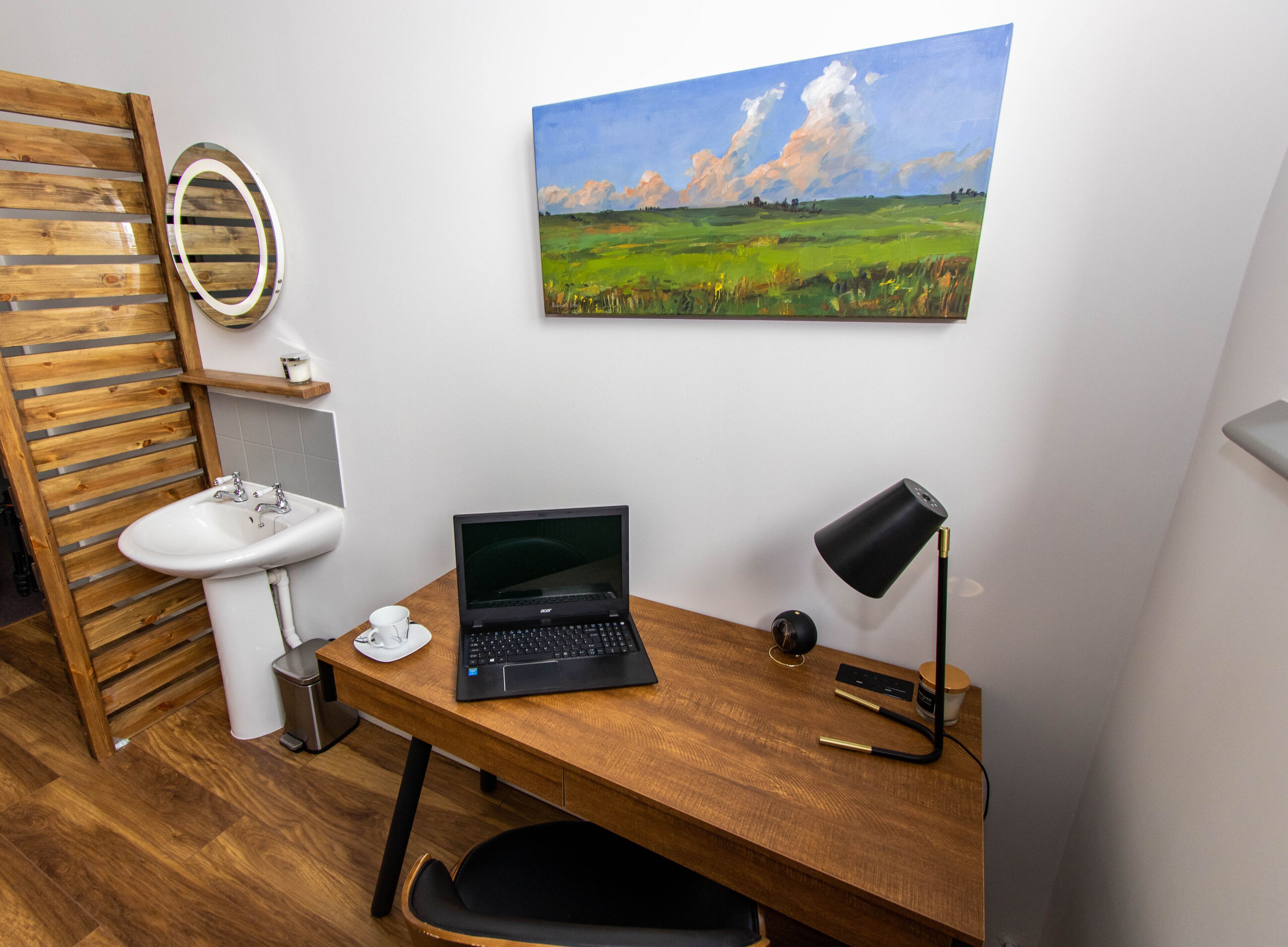 A room with a wooden desk, a laptop, a black desk lamp, a white sink, a mirror, and a painting of a landscape.