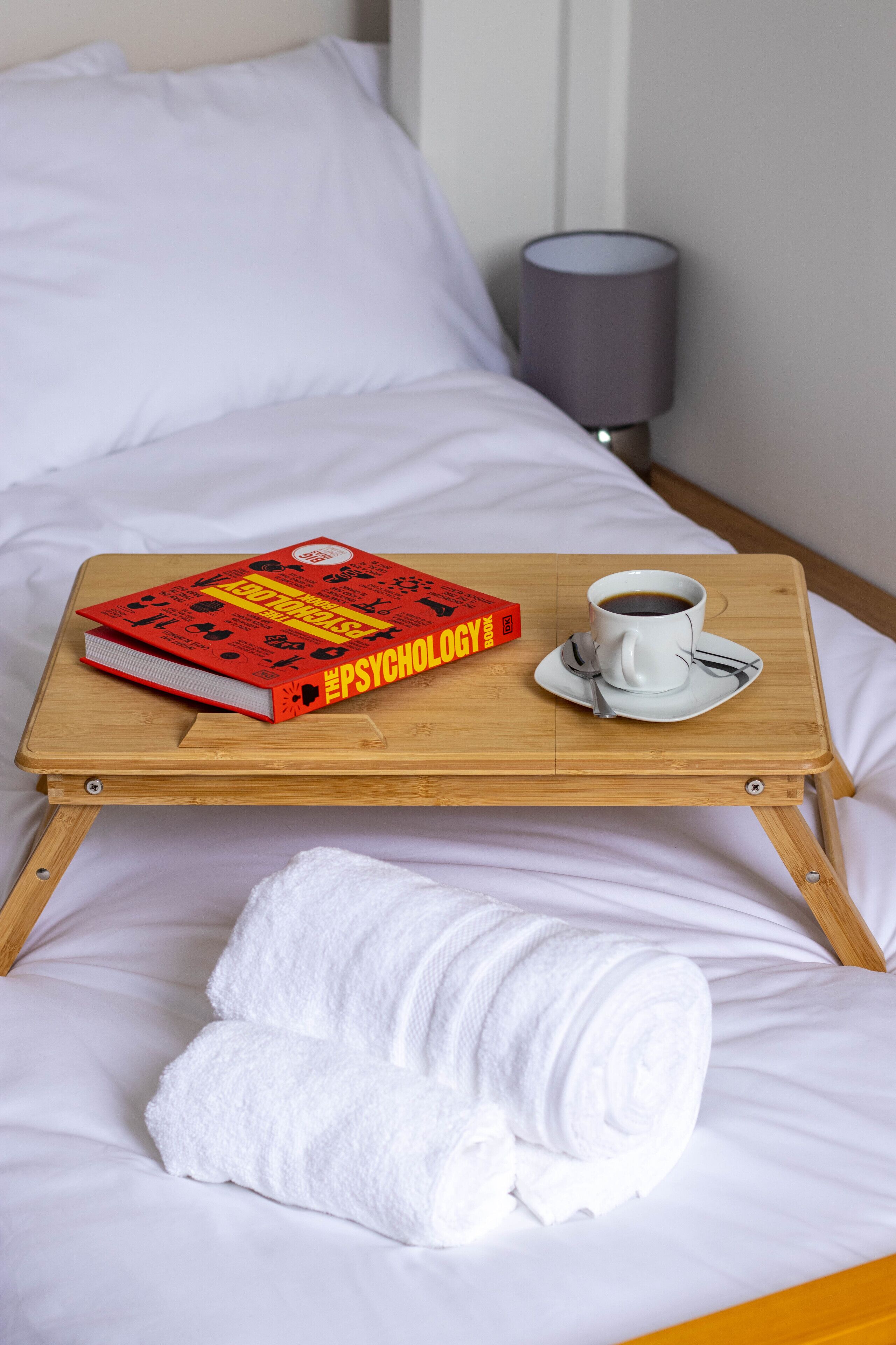 A bed with a wooden tray holding a book and a cup of coffee, a rolled-up towel, and a lamp in the background.