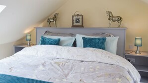 Een slaapkamer met een schuine plafond, een bed met bloemenbeddels, twee nachtkastjes met lampen en twee decoratieve paardenbeeldjes op het hoofdeinde.