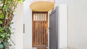 A narrow entrance with a wooden door and a grey gate, leading to a building with a white exterior and a curved roof. - Seaside villa sleeps 6, garden, air conditioning, garage (La Grande-Motte)