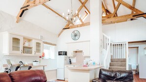 A spacious living area with a high ceiling, exposed wooden beams, a chandelier, a kitchen with white cabinets, and a staircase leading to an upper level.