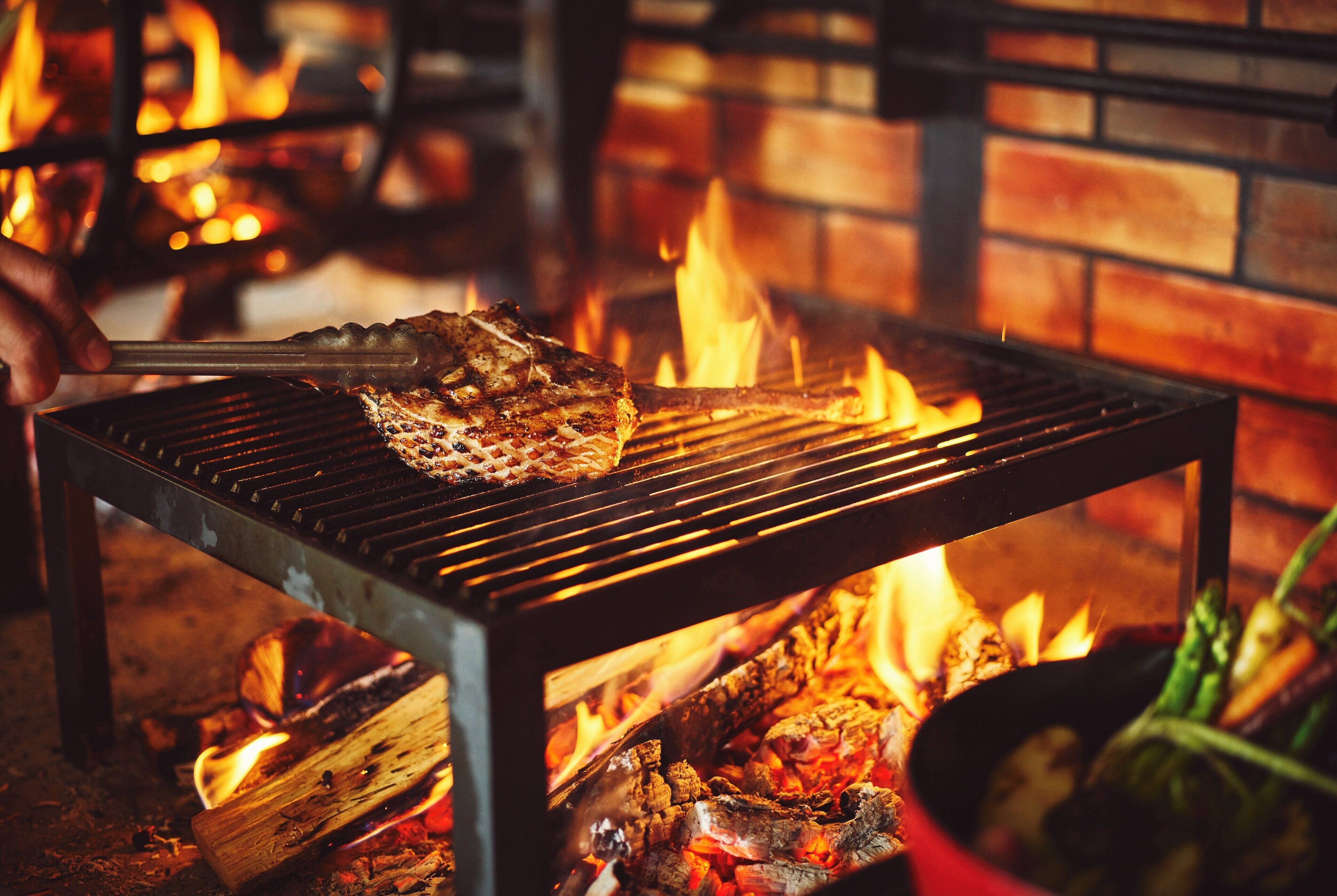 A grill with meat being cooked over an open flame, with a brick wall in the background.