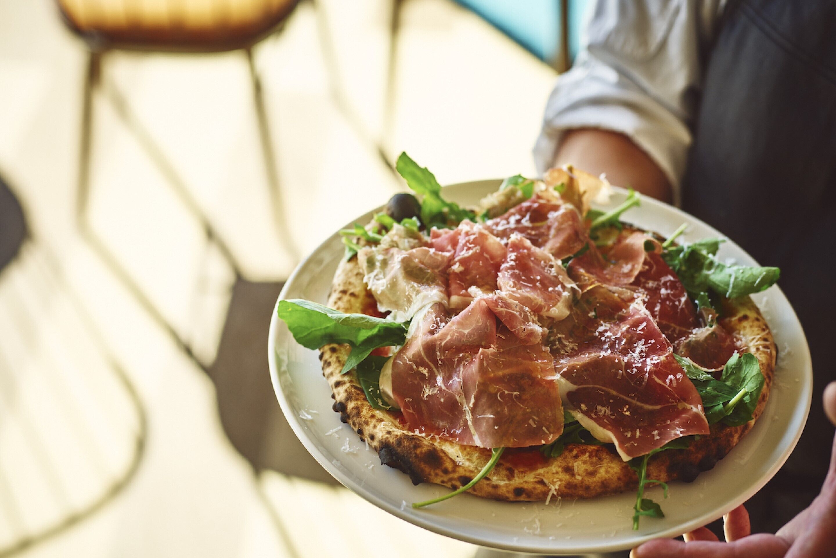 A person holding a plate with a pizza topped with greens and cured meat.