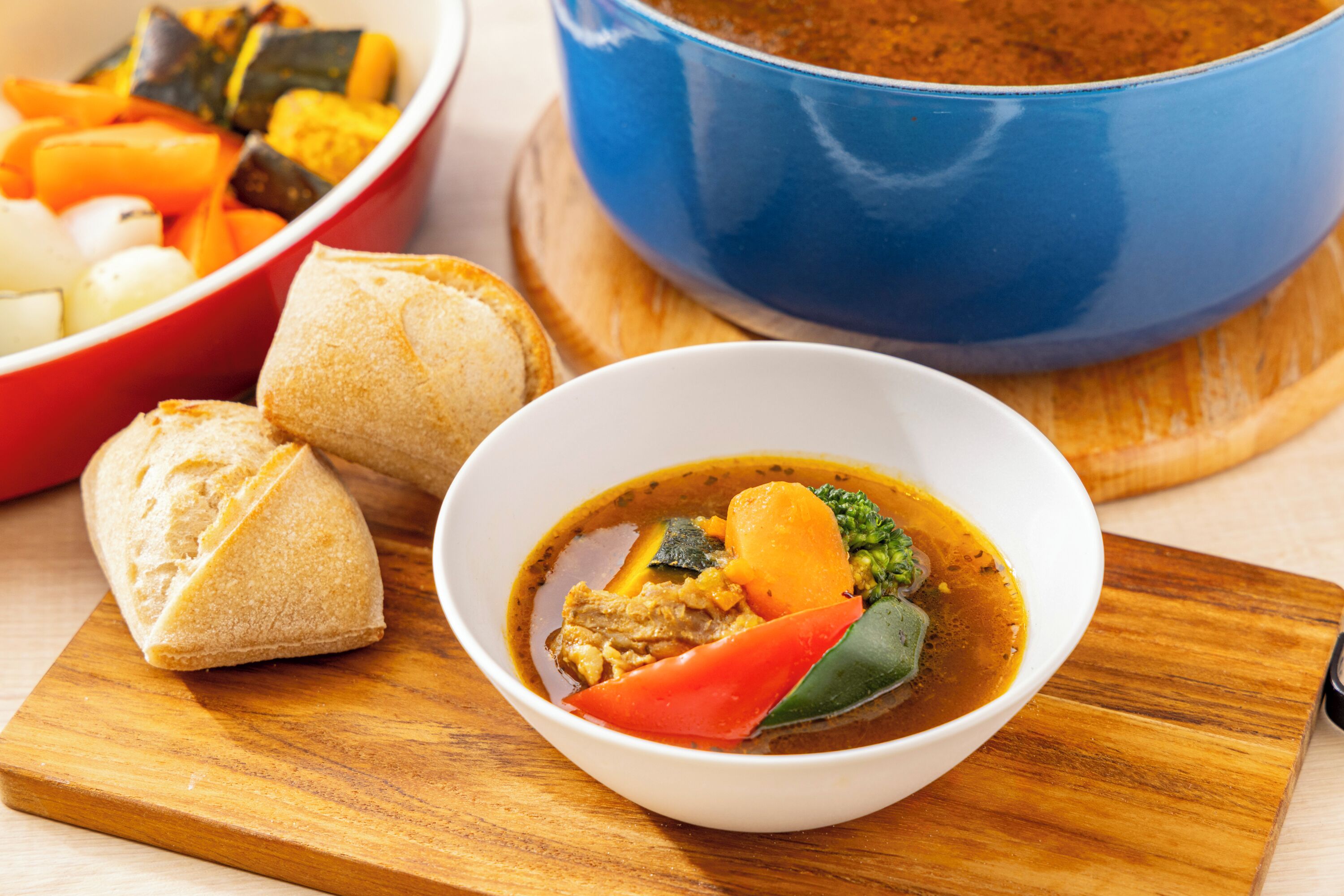 A bowl of soup with vegetables and meat, accompanied by bread on a wooden board.