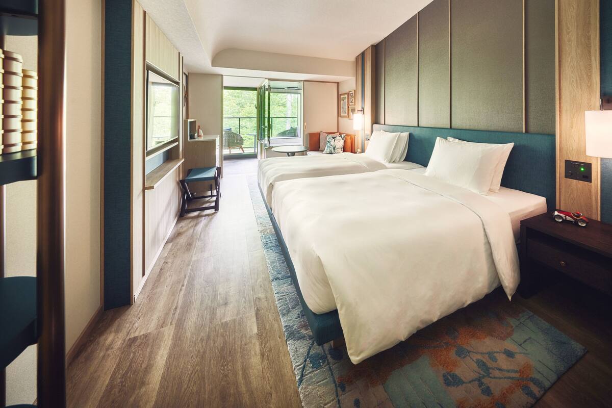 A hotel room with two beds, a balcony, a desk, and a view of greenery.