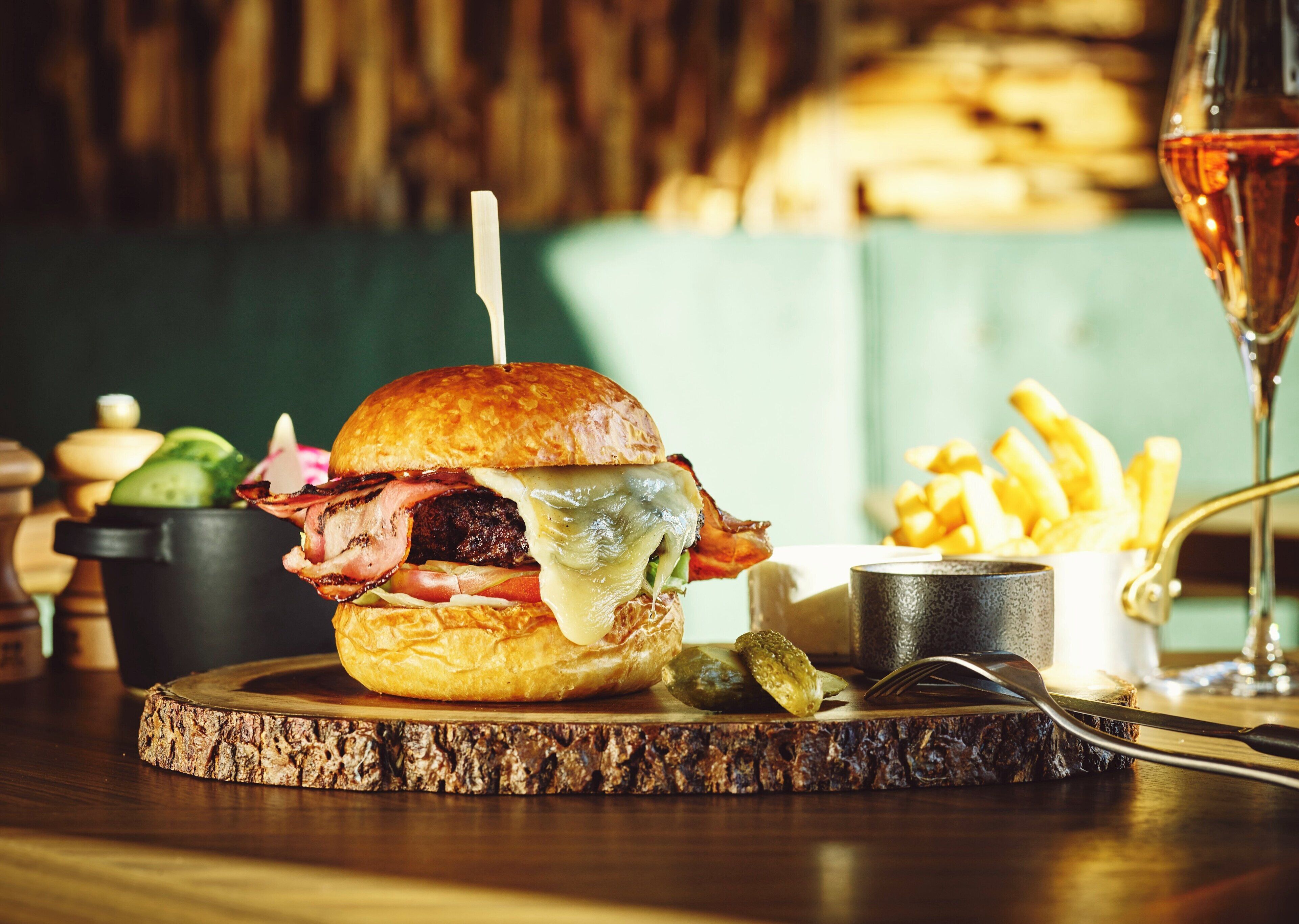 A burger with pickles and bacon, a glass of red wine, and a side of fries on a wooden board.