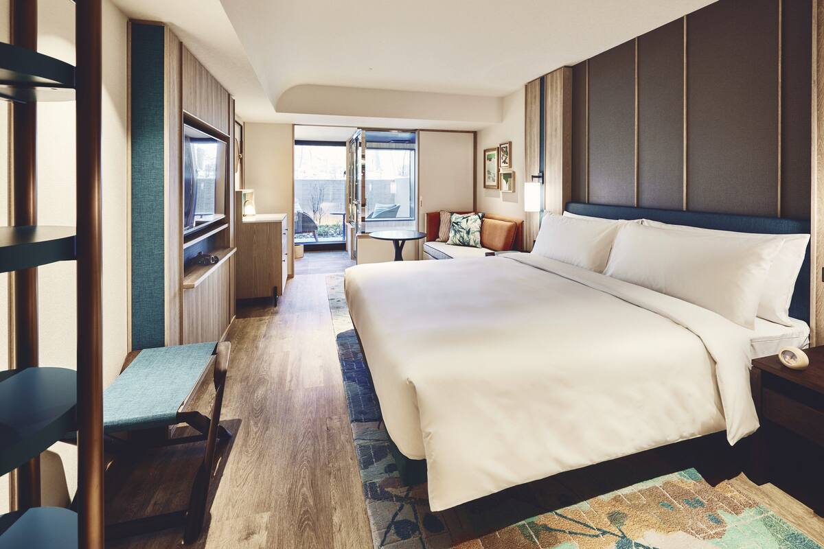 A hotel room with a large bed, a desk, a chair, a TV, and a view of the outside.
