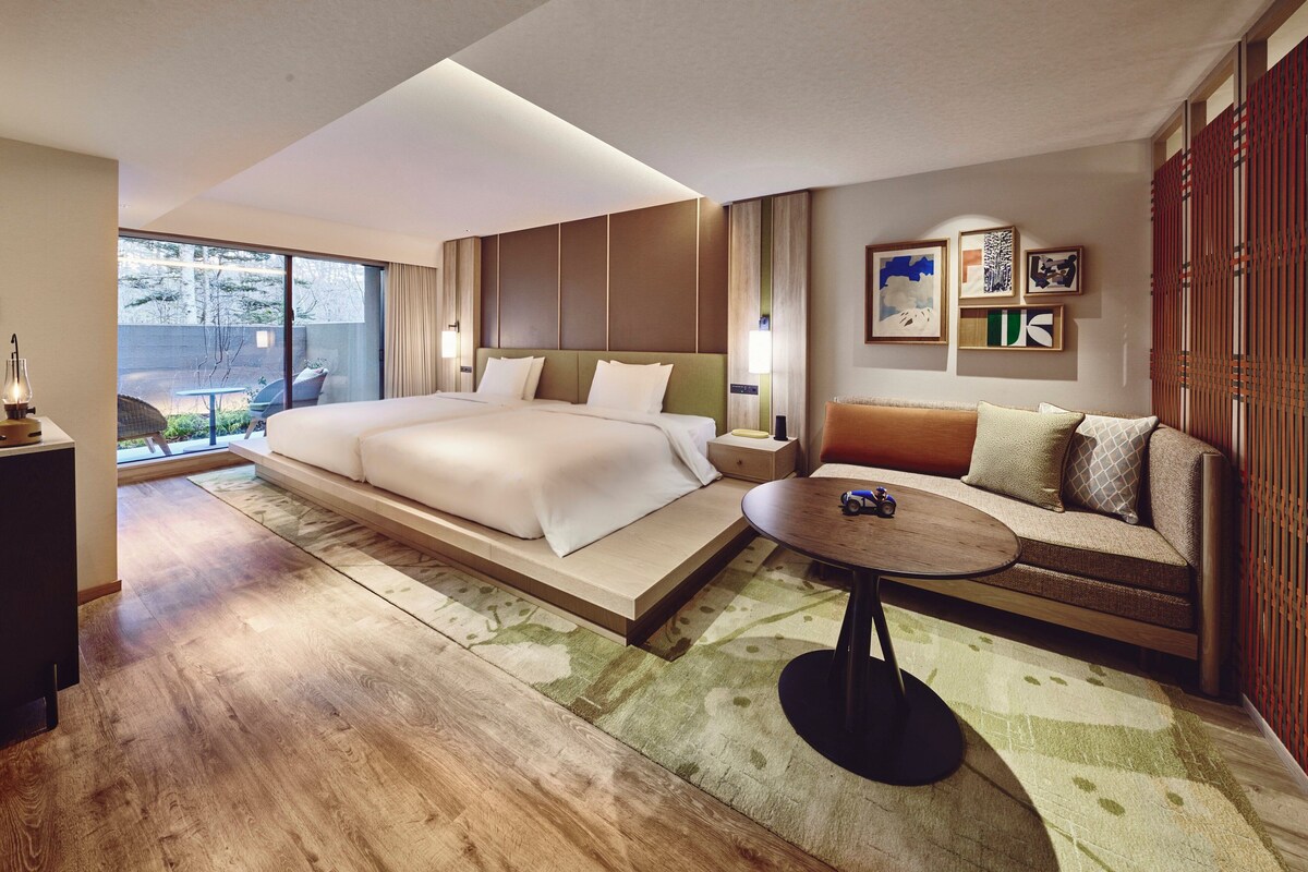 A modern hotel room with a large bed, a sofa, a small table, and a view of the outdoors.