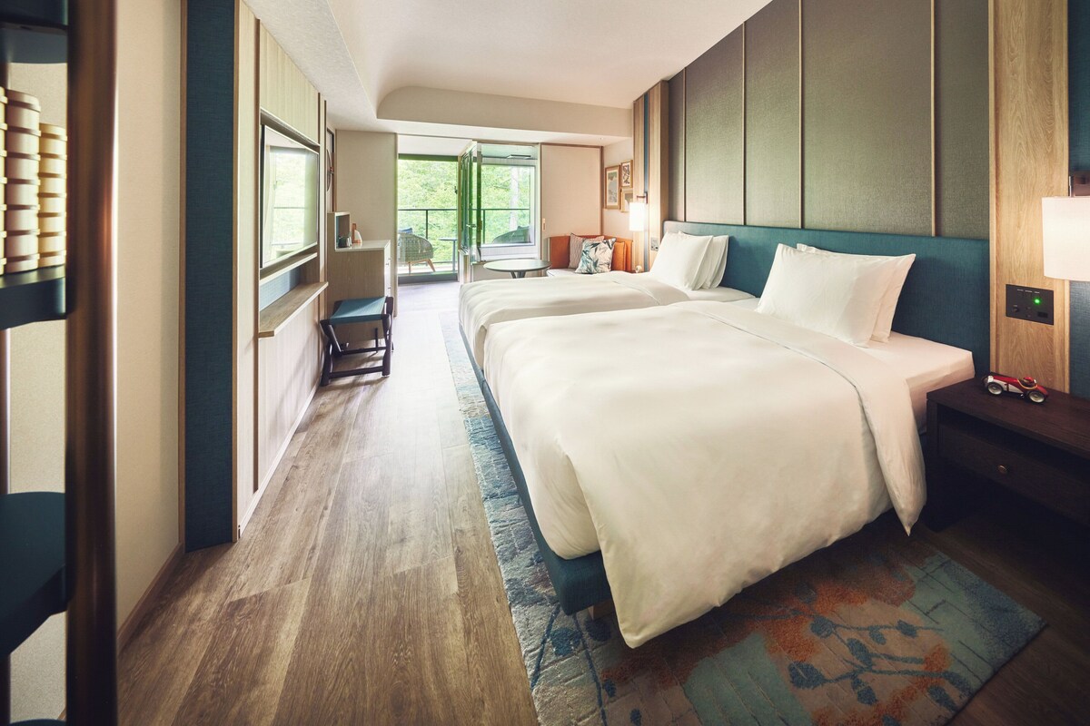 A hotel room with two beds, a balcony, a desk, and a view of greenery.