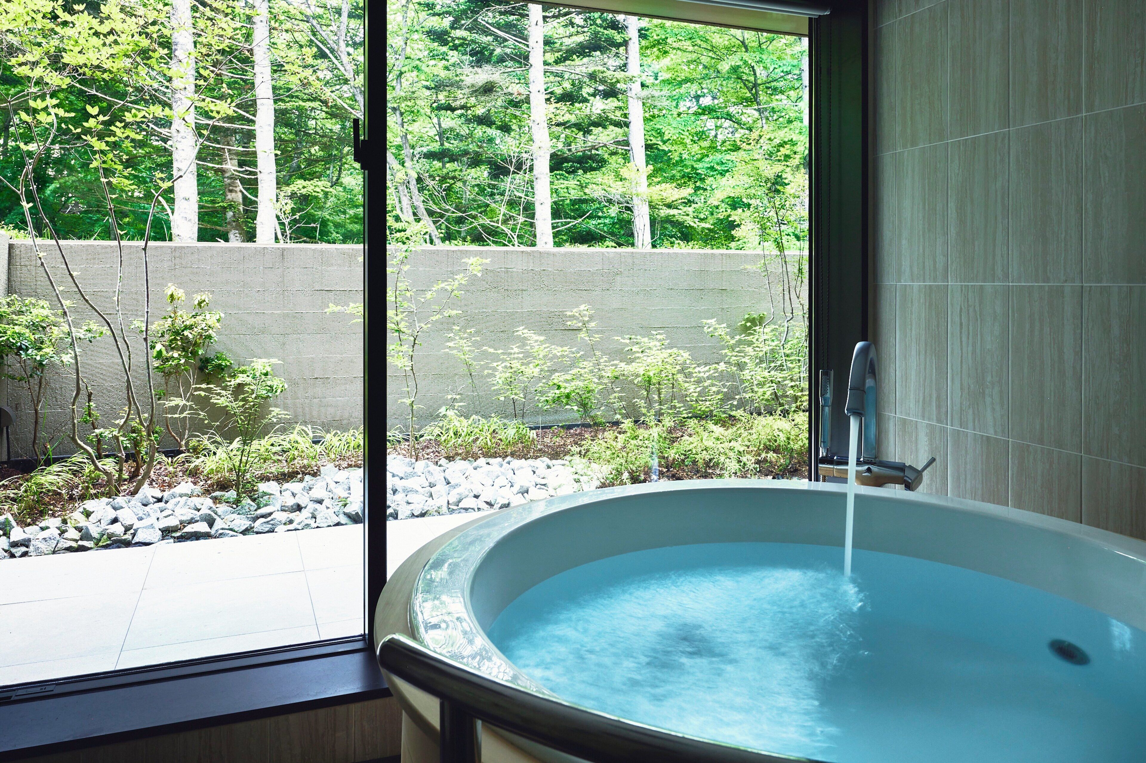 A modern bathroom with a large bathtub, a window with a view of a garden and trees, and a concrete wall.
