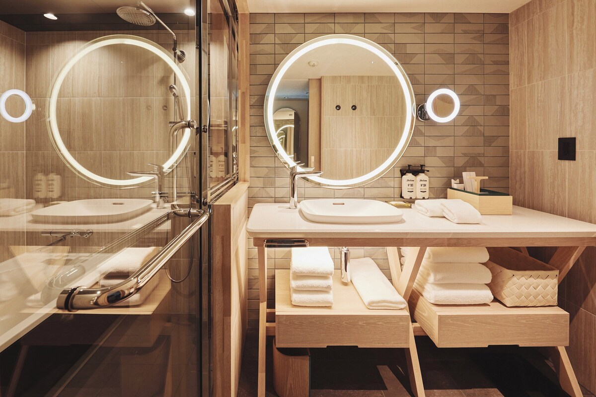 A modern bathroom with two sinks, each with a round mirror and under-cabinet lighting. There are towels and toiletries on the counter.
