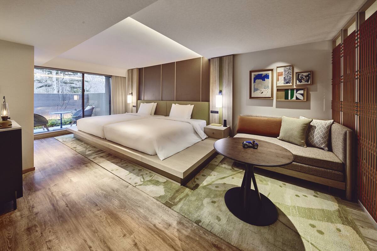 A modern hotel room with a large bed, a sofa, a small table, and a view of the outdoors.