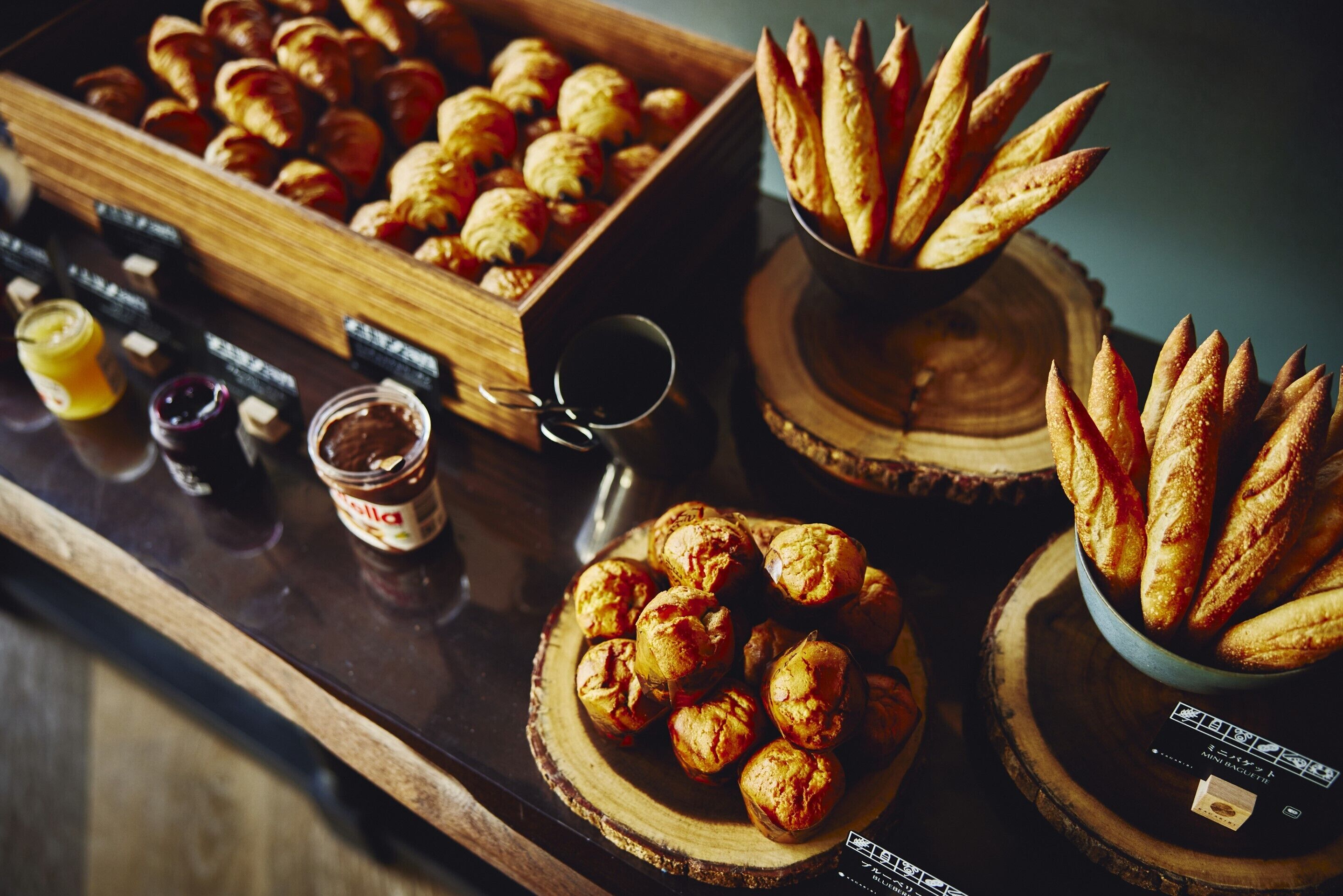 Une présentation de viennoiseries et de pains sur des planches en bois, avec des pots de confitures et une tasse de café.
