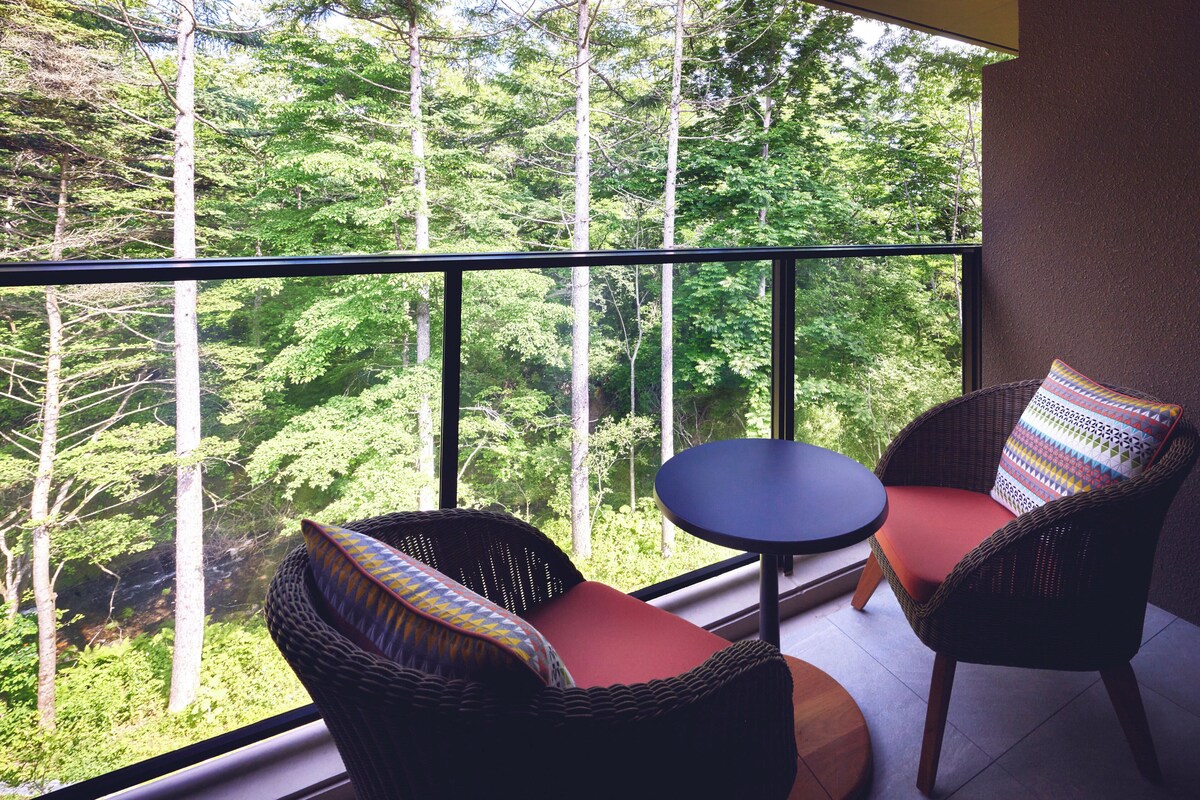 A balcony with wicker chairs and a small table, overlooking a forested area.