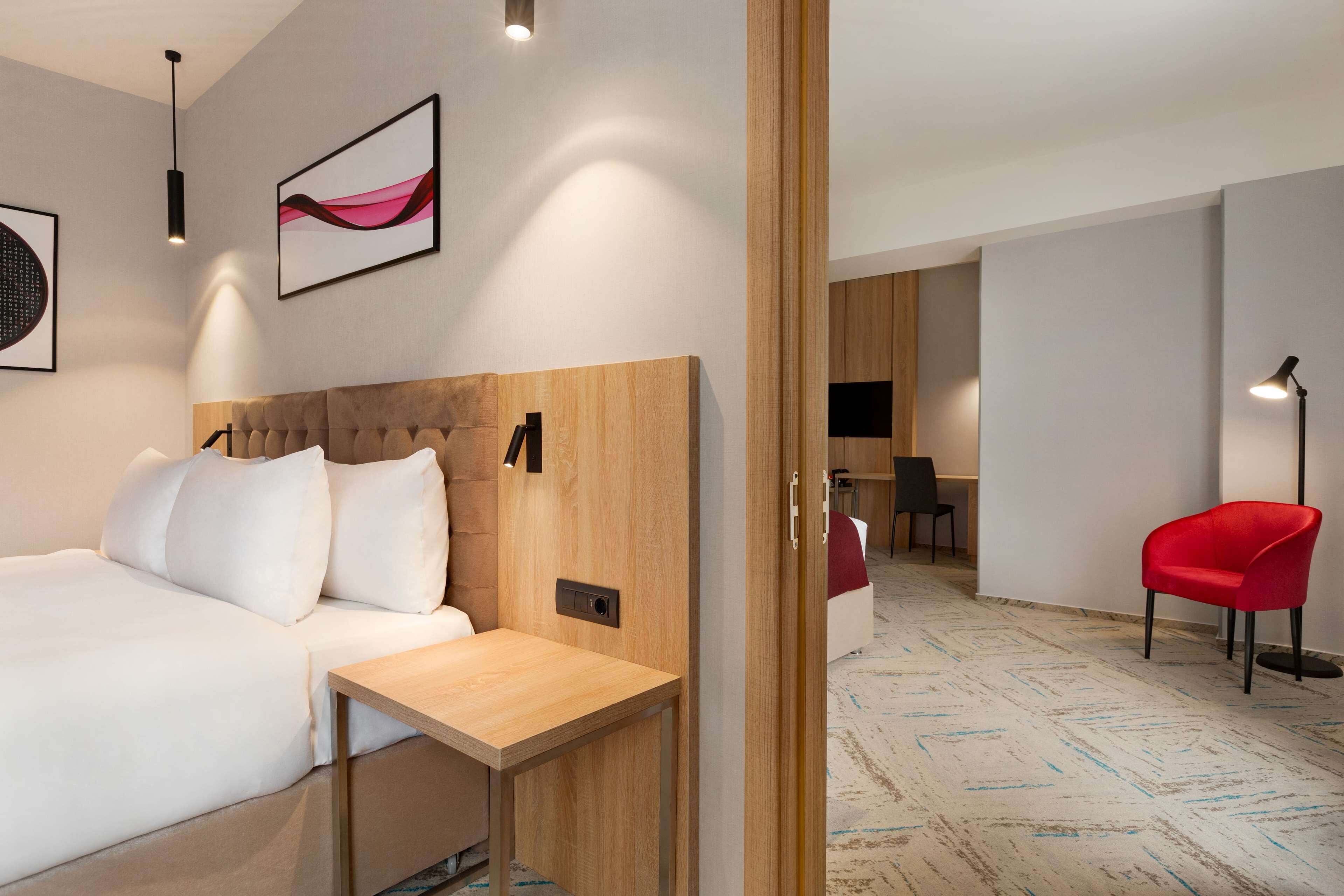 Une chambre d’hôtel moderne avec une tête de lit en bois, une table de chevet en bois, un lit blanc, une chaise rouge et un bureau avec une chaise.