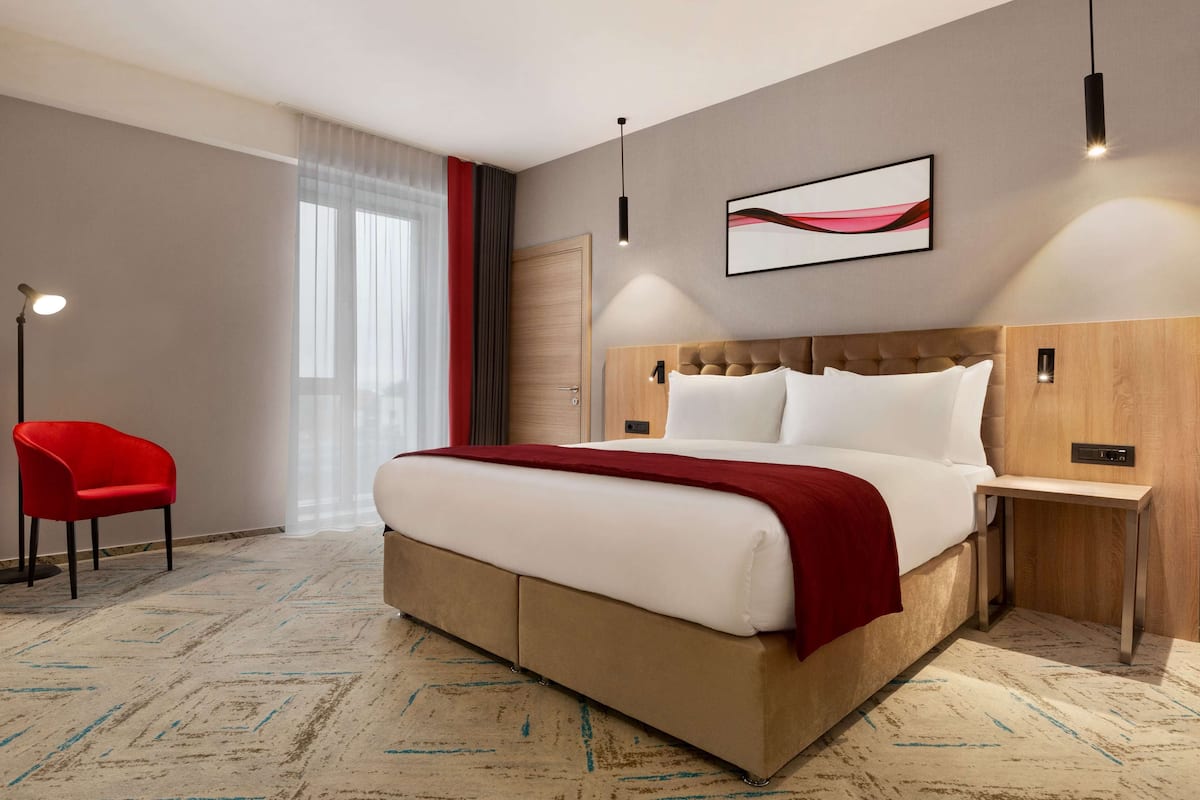 Hotelkamer met een groot bed, een rode stoel, een houten hoofdeinde, een moderne hanglamp en een kunstwerk in een lijst aan de muur.