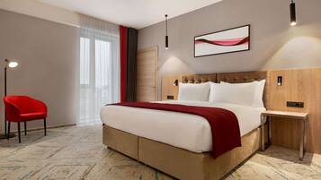 A hotel room with a large bed, a red chair, a wooden headboard, a modern pendant light, and a framed artwork on the wall.