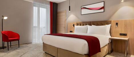 Habitación de hotel con una cama grande, una silla roja, un cabecero de madera, una lámpara colgante moderna y una obra de arte enmarcada en la pared.