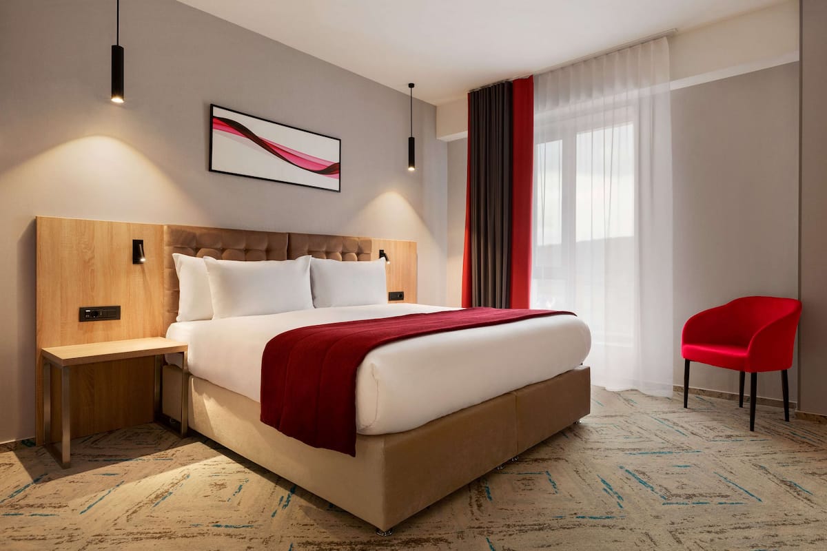 Een moderne hotelkamer met een groot bed, een houten hoofdbord, een rode stoel en een groot raam met dunne gordijnen.