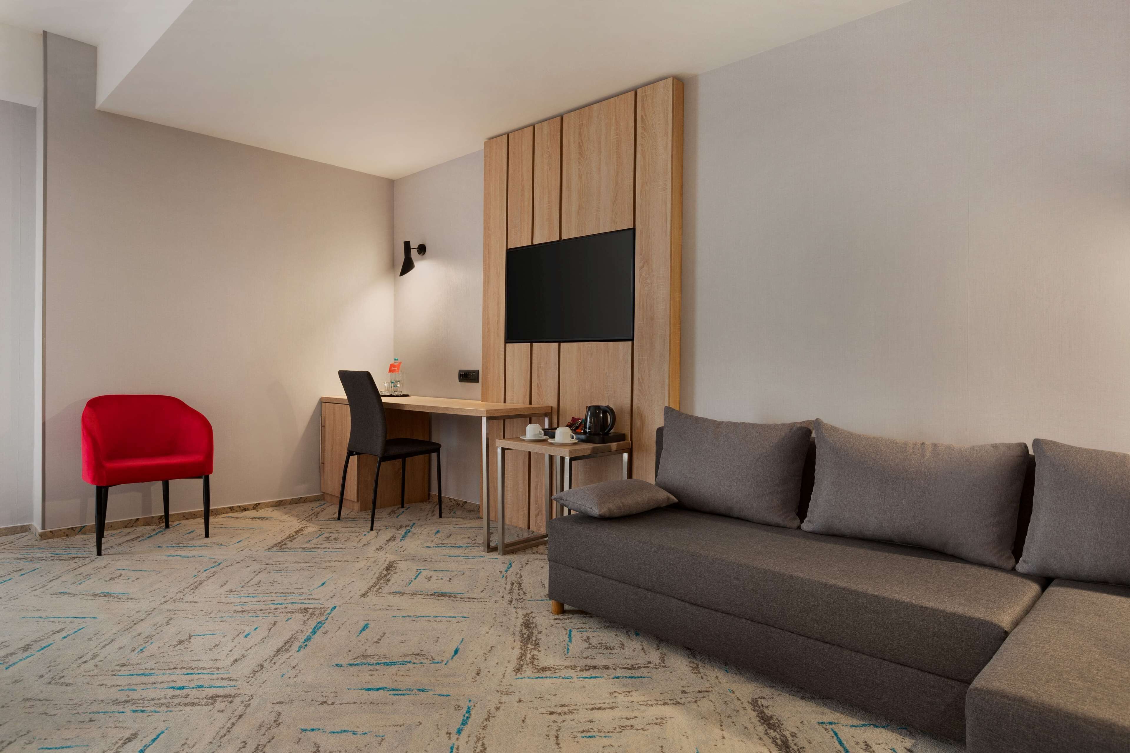 Una habitación de hotel moderna con un sofá gris, una silla roja, un escritorio de madera y un televisor de pantalla plana, además de una alfombra estampada.