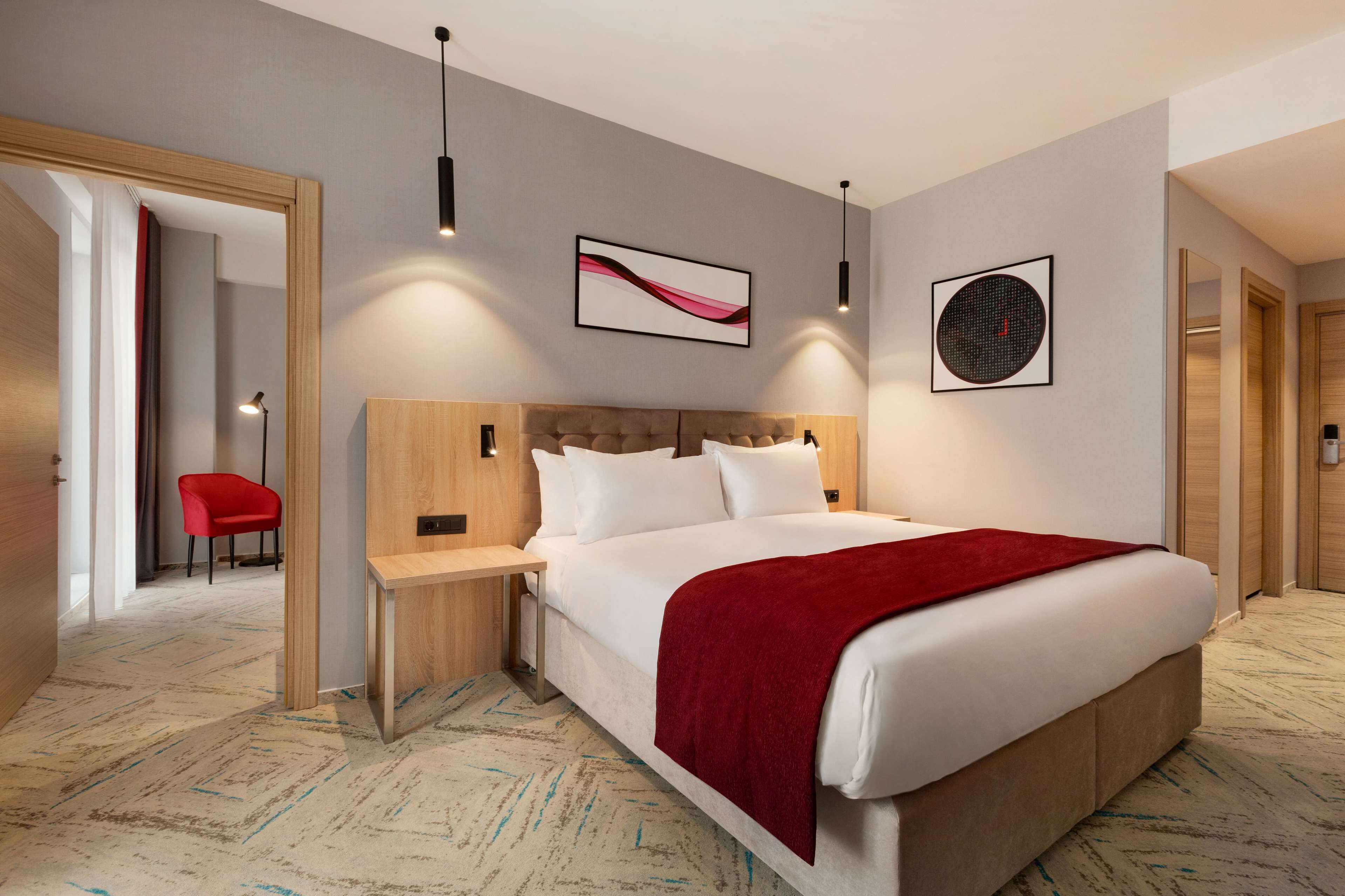 A modern hotel room with a large bed, a wooden headboard, a red throw blanket, and two wall-mounted artwork pieces.
