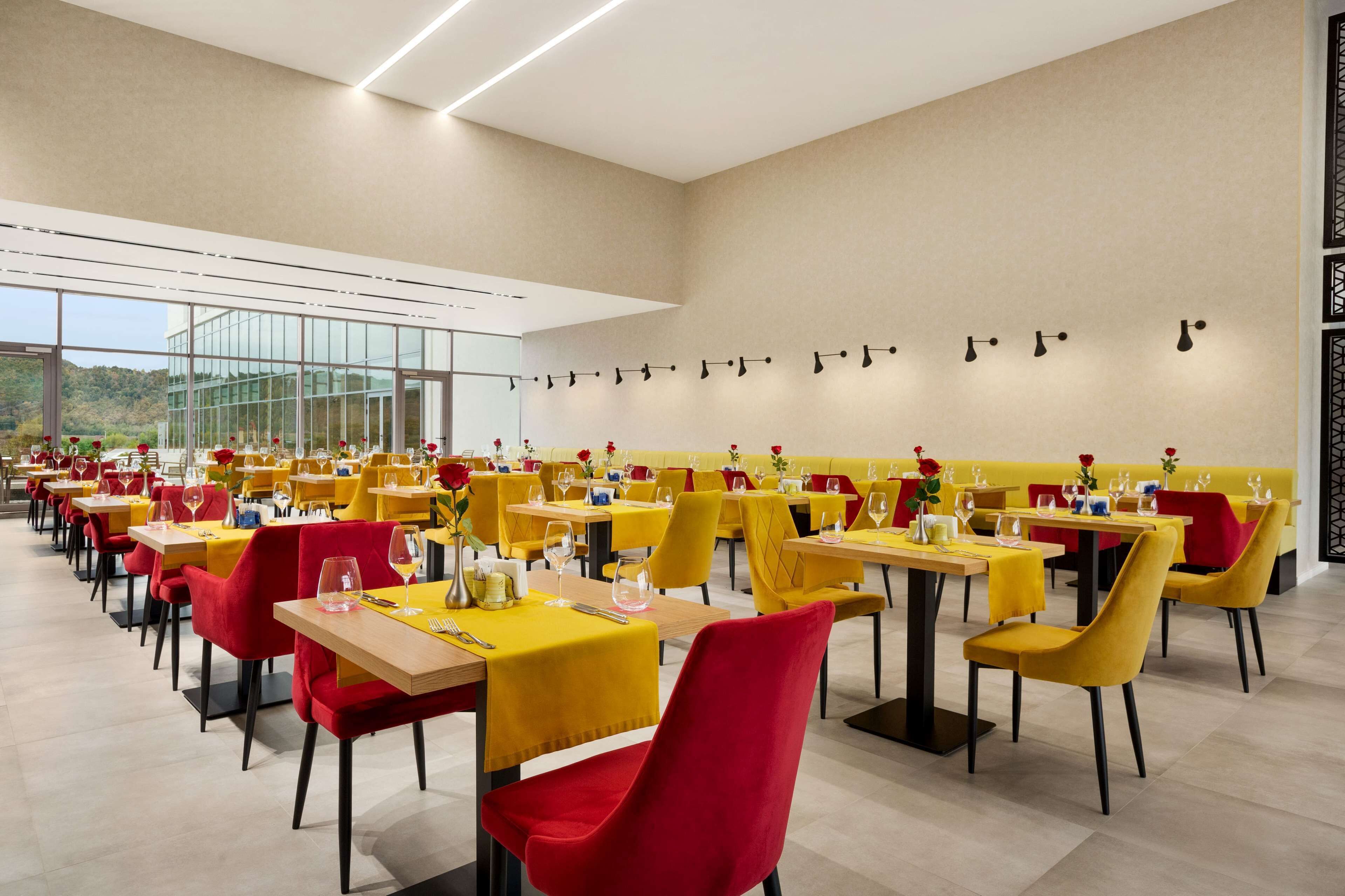 A modern dining area with yellow tables and red chairs, large windows, and a view of the outdoors.