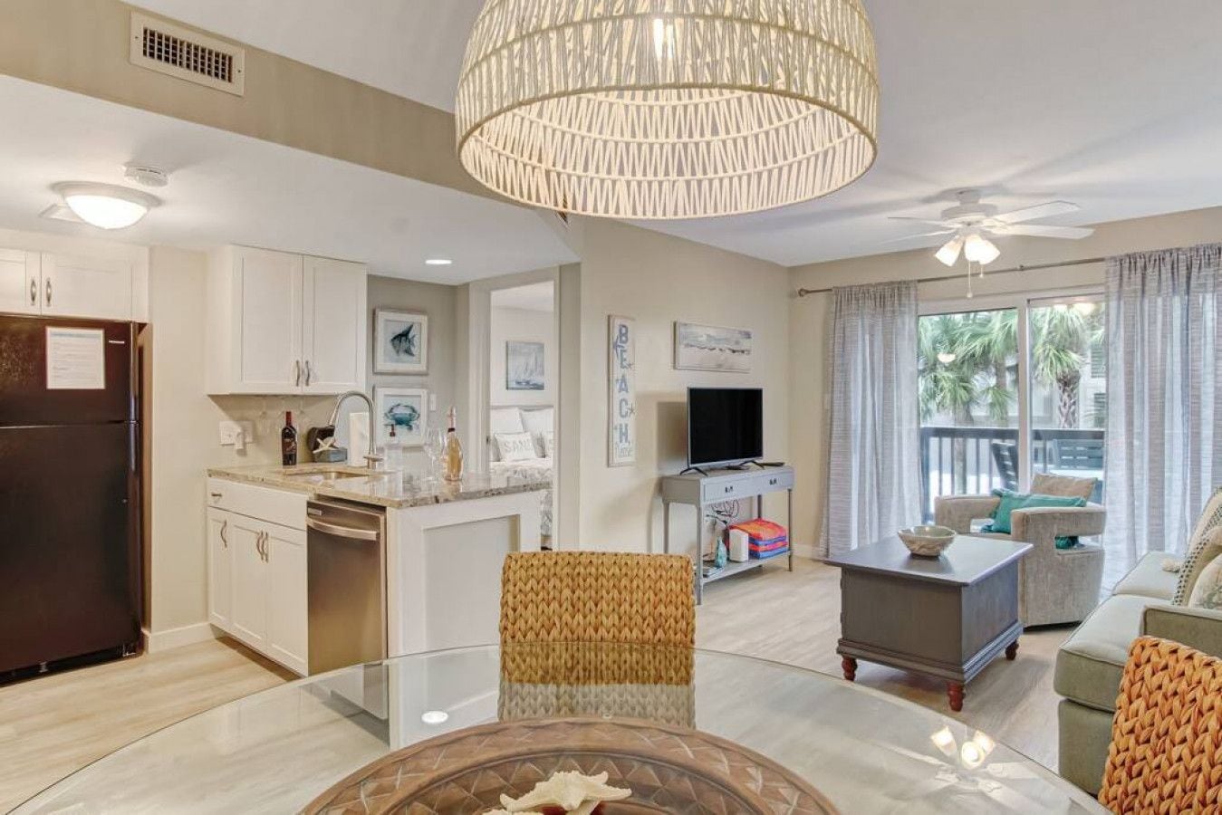 A modern living room with a glass coffee table, a dining area with a white table, and a kitchen with white cabinets and a dark refrigerator.