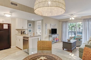 A modern living room with a glass coffee table, a dining area with a white table, and a kitchen with white cabinets and a dark refrigerator. - Oceanview Sand Dollar Villas Condo | Balcony | Pool (Fernandina Beach)