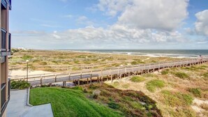 Een kustgezicht met een houten vogelpromenade die naar het strand leidt, een grasveld op de voorgrond en een gebouw met een balkon met uitzicht op het tafereel.