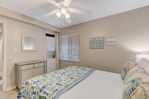 A bedroom with a bed, a ceiling fan, a window with blinds, a dresser, and a wall-mounted sign.