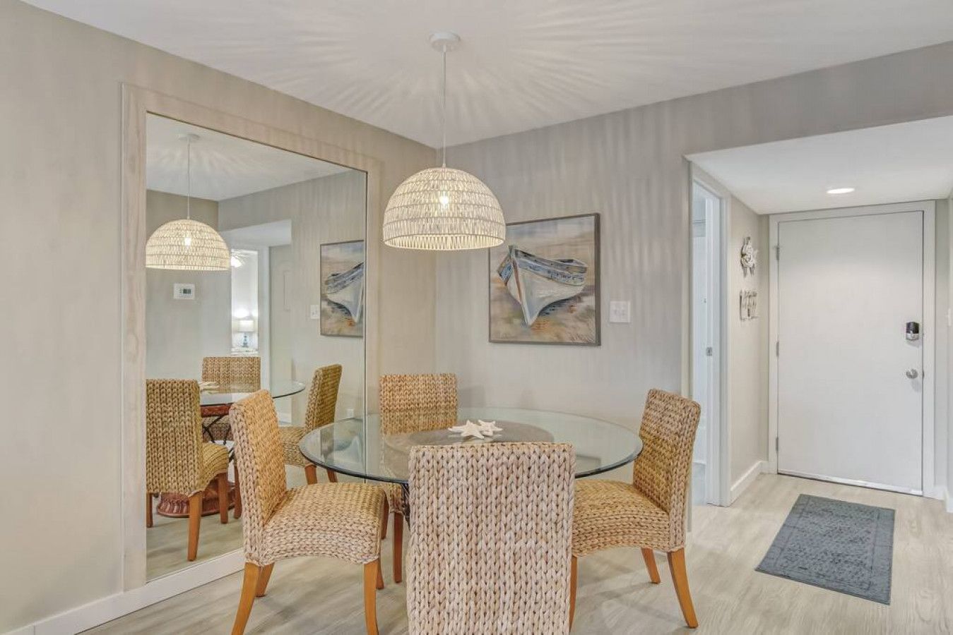 A dining area with a glass table surrounded by woven chairs, a large mirror, and two pendant lights.