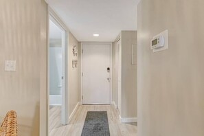 A hallway with a light-colored floor, a gray rug, a white door, and a thermostat on the wall.