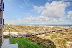 A coastal view with a wooden boardwalk leading to the beach, a grassy area in the foreground, and a building with a balcony overlooking the scene. - Oceanview Sand Dollar Villas Condo | Balcony | Pool (Fernandina Beach)