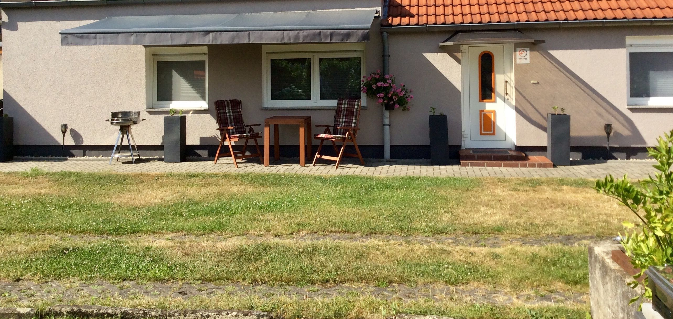 Ein Haus mit einer Terrasse, auf der sich ein Grill, zwei Stühle und ein Tisch befinden.