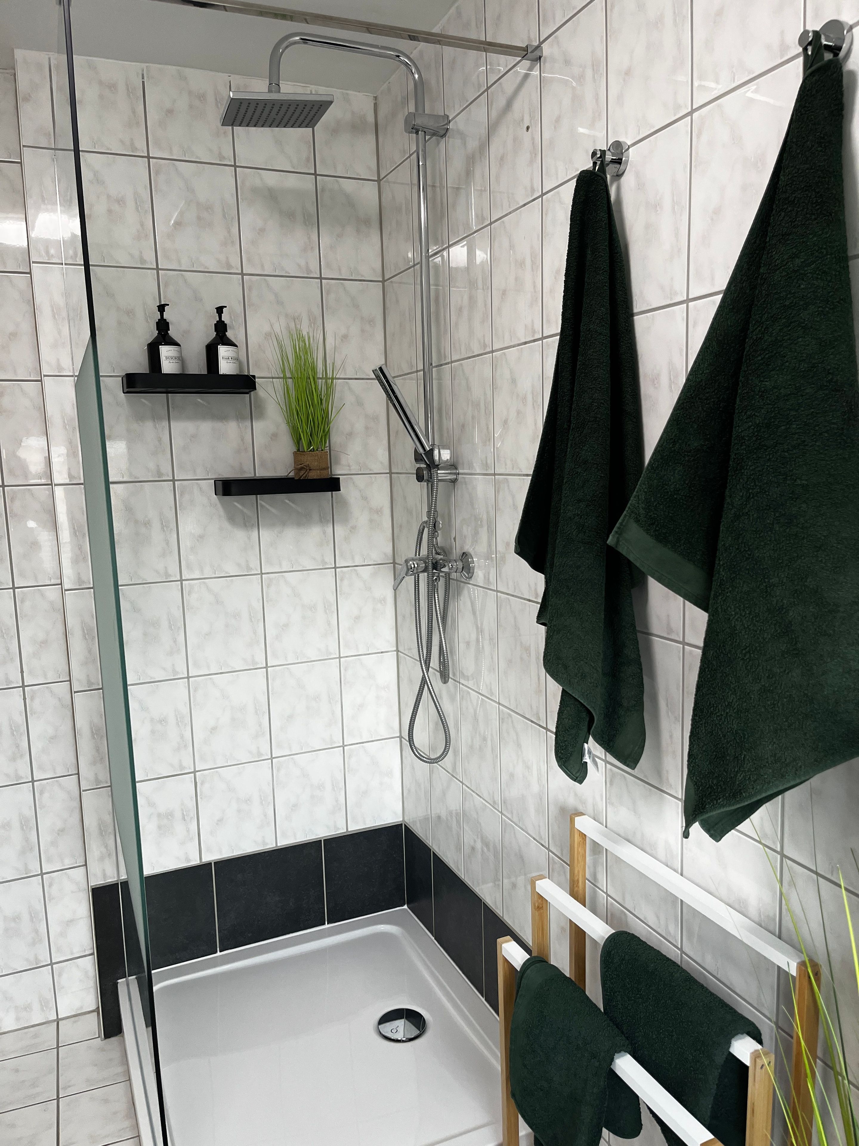 Ein modernes Badezimmer mit Duschkabine aus Glas, schwarz-weißen Fliesen, einer kleinen Ablage für Pflegeprodukte und einem grünen Handtuchhalter.