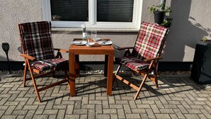Ein Sitzbereich im Freien mit zwei Holzstühlen und einem Tisch, vor einem Gebäude mit einem Fenster und Pflanzen in Töpfen.