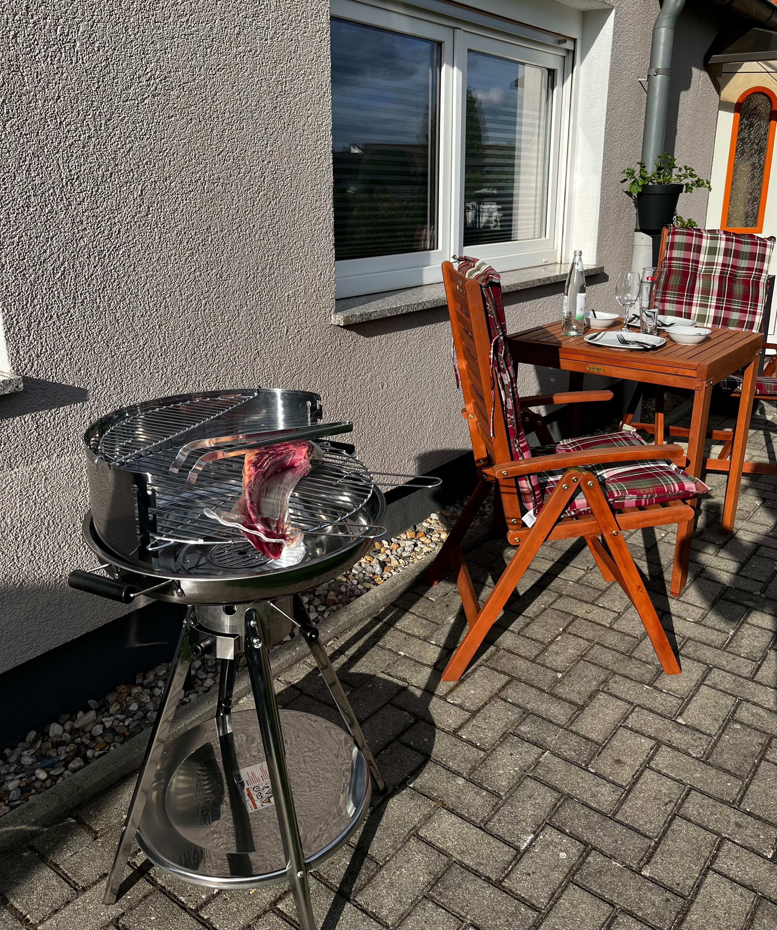 Eine Terrasse im Freien mit einem runden Tisch und Stühlen, einem Grill und einem Fenster mit Jalousien.