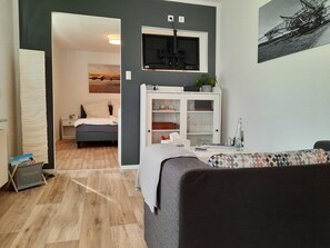 A modern living room with a grey sofa, a wooden floor, a TV mounted on the wall, and a cabinet with glass doors. - Vacation apartment Paula (Senftenberg)