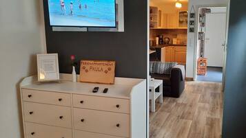Een moderne woonkamer met een televisie waarop een strandscène te zien is, een wit bergkastje met lades, een kleine tafel met een vaas en een bord, en een houten vloer.