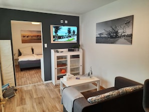 A modern living room with a sofa, a small table, a TV, a bookshelf, and a bed in the background. - Vacation apartment Paula (Senftenberg)