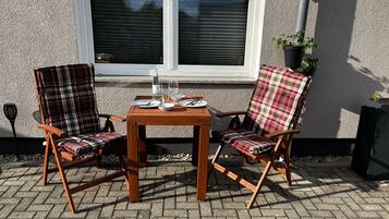 Een buitenruimte met twee houten stoelen en een tafel, voor een gebouw met een raam en bloempotten.