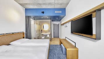 Una habitación de hotel moderna con dos camas, un televisor grande, un baño con espejo y lavamanos, y una pequeña zona de estar.