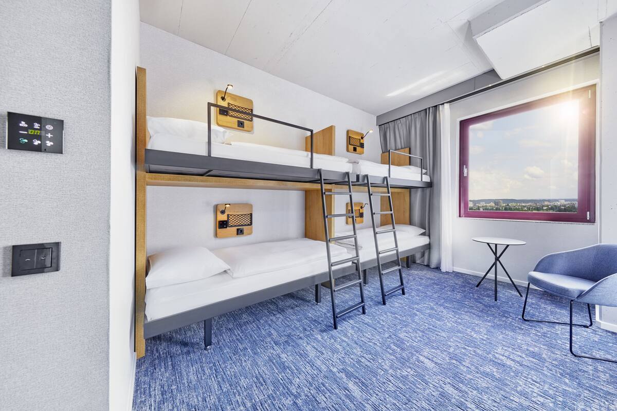 Une chambre d’hôtel avec des lits superposés, une petite table, une chaise bleue et une fenêtre donnant sur la ville.