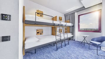 A hotel room with bunk beds, a small table, a blue chair, and a window with a city view.