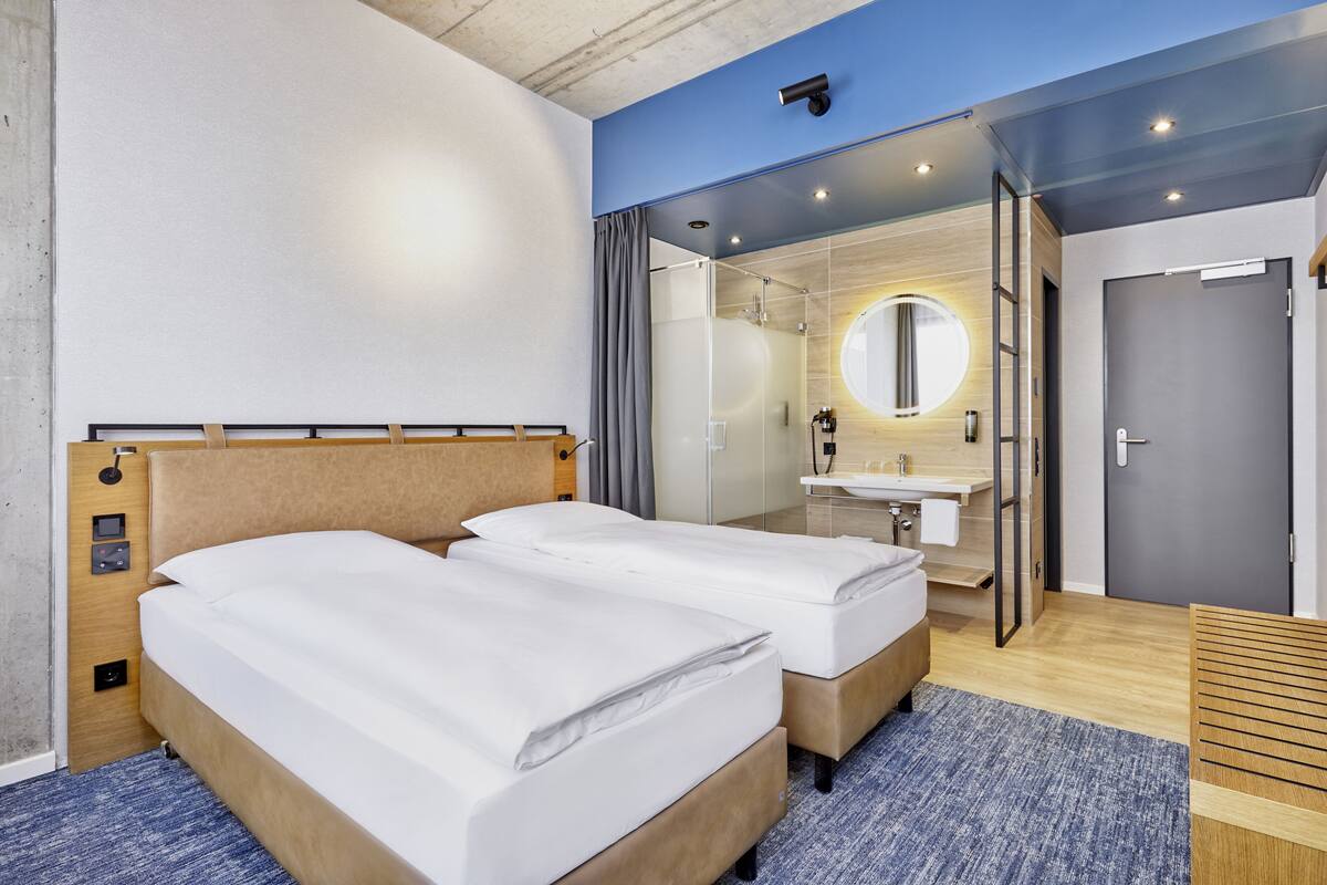 Une chambre d’hôtel moderne avec deux lits, une salle de bain dotée d’un miroir et d’un lavabo, et une vue sur la ville.