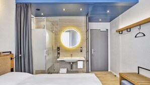 A modern hotel room with a large bed, a bathroom with a glass shower enclosure, a round mirror with a light, and a wooden bench. - Sunday Hotel Düsseldorf Seestern (Düsseldorf)