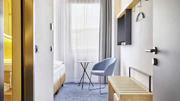 Quarto de hotel com uma cama, uma mesinha, uma cadeira, televisão na parede e janela com cortinas.