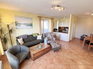 A living room with a sofa, armchair, coffee table, and a dining area with a table and chairs. There is a kitchen area with a counter and appliances visible through an open door.