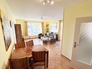 A living room with a dining table, chairs, a sofa, and a coffee table. There is a door on the right and a window with curtains.